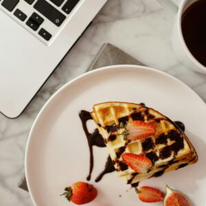 Top view of a waffle with chocolate and strawberries next to a laptop and coffee.